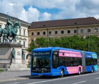 Ein knallblauer Elektro-Bus mit pinkem Element fährt vor historischen Gebäuden in München entlang.