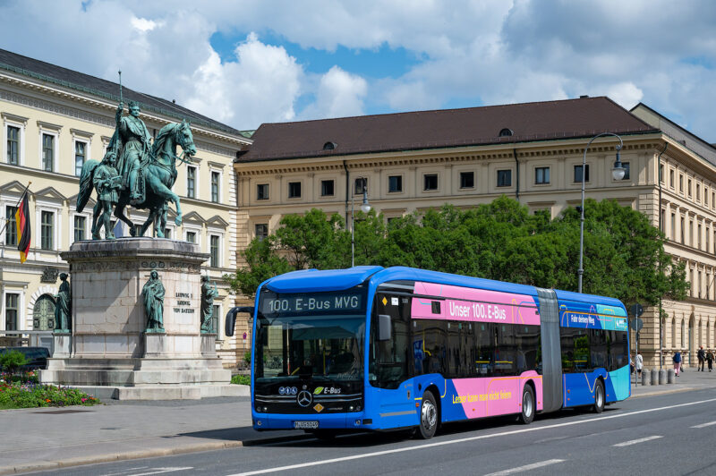 Ein knallblauer Elektro-Bus mit pinkem Element fährt vor historischen Gebäuden in München entlang.