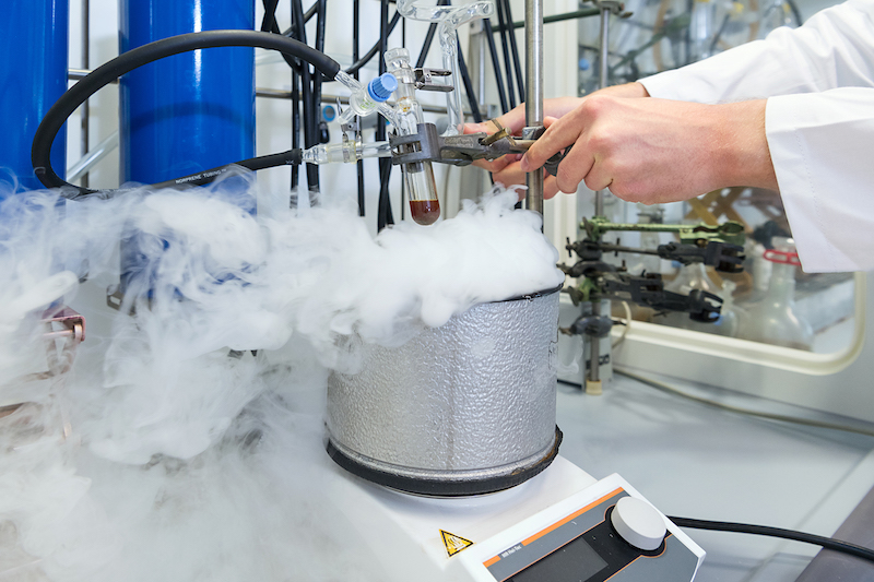 Im chemischen Labor steht ein dampfender Topf auf einem Kochfeld. Ein wissenschaftler experimentiert mit einer dunklen Flüssigkeit in einem Reagenzglas.