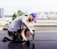 Das Foto zeigt einen Monteur mit einem Akku-Schrauber, der ein PV-Modul befestigt. Er trägt eine lila Kappe, ein fliederfarbenes T-Shirt sowie eine schwarze Arbeitshose. Er hockt neben dem Modul und ist von der Seite fotografiert.