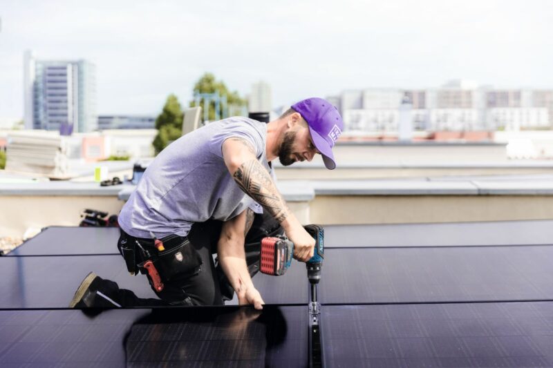 Das Foto zeigt einen Monteur mit einem Akku-Schrauber, der ein PV-Modul befestigt. Er trägt eine lila Kappe, ein fliederfarbenes T-Shirt sowie eine schwarze Arbeitshose. Er hockt neben dem Modul und ist von der Seite fotografiert.