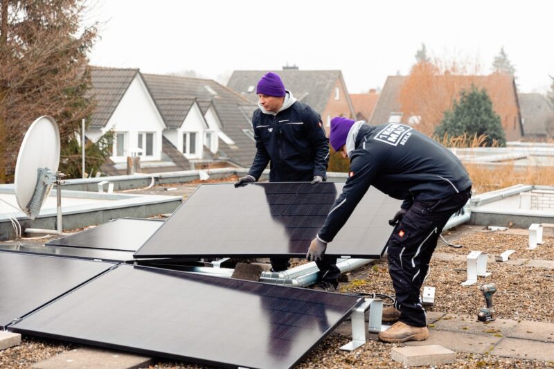 Zwei Männer mit Jacken auf denen 1Komma5° steht, montieren PV-Module auf einem Dach.