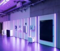 Ausstellungsfläche mit verschiedenen Energietechnik-Komponenten an einer beleuchteten Wand. Zu sehen sind unter anderem Geräte für Photovoltaik, Batteriespeicher, Wärmepumpen und Energiemanagement. Violette LED-Lichtstreifen und ein modernes Showroom-Design prägen die Szene.