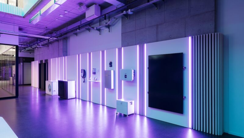 Ausstellungsfläche mit verschiedenen Energietechnik-Komponenten an einer beleuchteten Wand. Zu sehen sind unter anderem Geräte für Photovoltaik, Batteriespeicher, Wärmepumpen und Energiemanagement. Violette LED-Lichtstreifen und ein modernes Showroom-Design prägen die Szene.