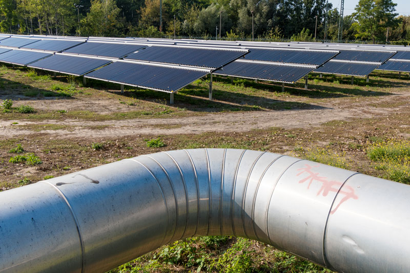 Symbolbild für BEW-Förderung: Vordergrund Fernwärmerohr, Hintergrund Solarthermieanlage auf Freifläche