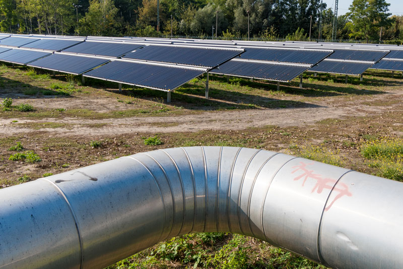 Symbolbild für BEW-Förderung: Vordergrund Fernwärmerohr, Hintergrund Solarthermieanlage auf Freifläche