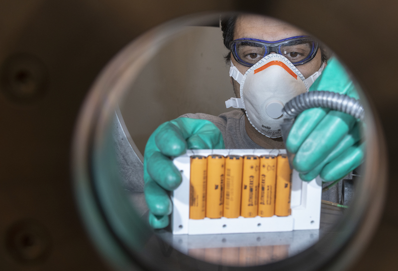 Blick durch ein Bullauge auf Mann in Laborkleidung mit Batterien in der Hand.