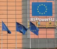 Die Flagge der Europäischen Union vor einem Gebäude der EU-Kommission. Die Fassade schimmert bronzen. Darauf unter einer weiteren Flagge der EU der Schriftzug "REPowerEU". Abgebildet sind auch Handwerker, die den Schriftzug zu befestigen scheinen - sie sind ein Bild. Flagge, Schriftzug und Handwerker sind Teile eines Banners an der Fassade.