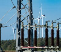 Durchsicht durch ein Umspannwerk auf einen Windpark. Im Vordergrund: Isolatoren, Strom- und Erdungsleitungen, Gittermasten.