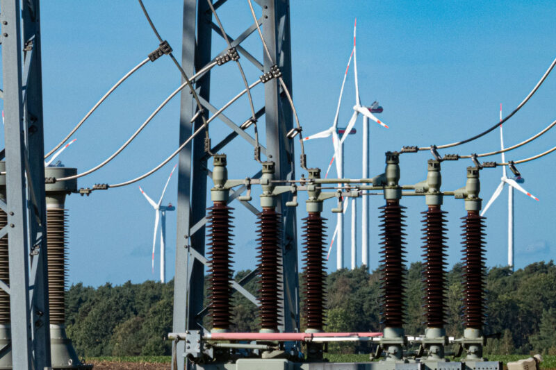 Durchsicht durch ein Umspannwerk auf einen Windpark. Im Vordergrund: Isolatoren, Strom- und Erdungsleitungen, Gittermasten.