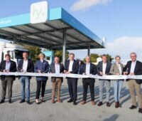 Mehrere Personen stehen draußen vor der Wasserstofftankstelle mit einer Schere vor einem hellen Band.