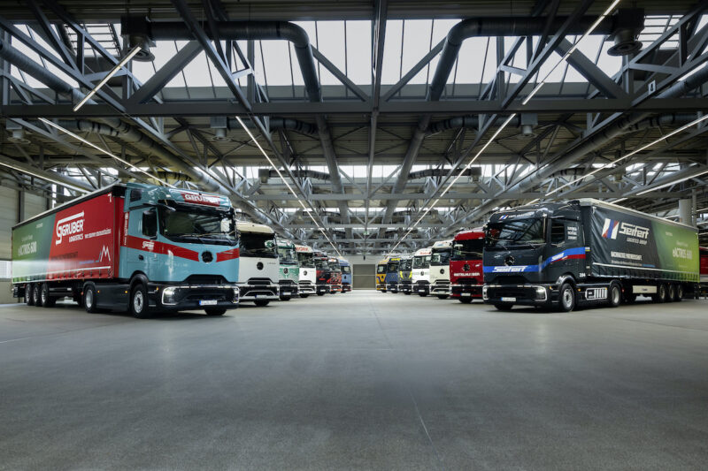 14 Trucks sind in einer riesigen Halle in zwei Reihen gegenüber stehend aufgereiht. Sie sind schräg nach vorn nebeneinander ausgerichtet.