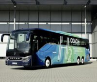 Ein Omnibus ist auf einem Betriebsgelände von schräg recht nach vorn aufgenommen. Er trägt auf blau bis türkisfarbenen Meeresfarben den Schriftzug H₂ Coach.