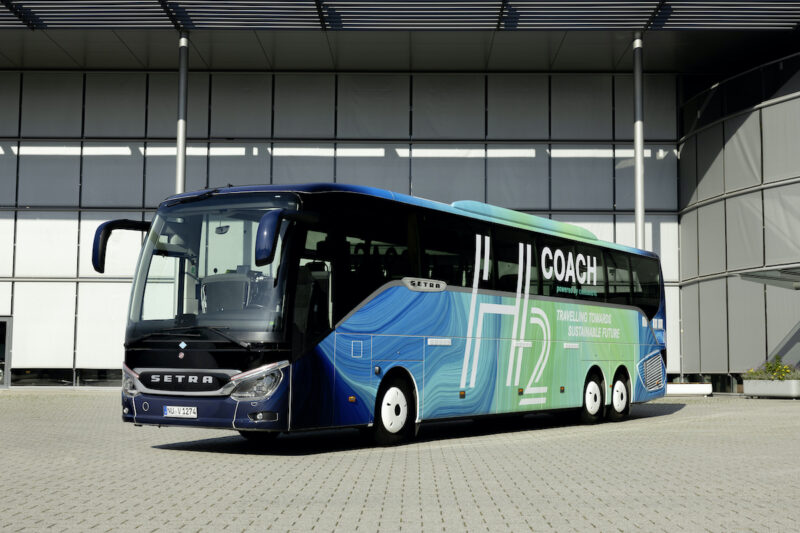 Ein Omnibus ist auf einem Betriebsgelände von schräg recht nach vorn aufgenommen. Er trägt auf blau bis türkisfarbenen Meeresfarben den Schriftzug H₂ Coach.