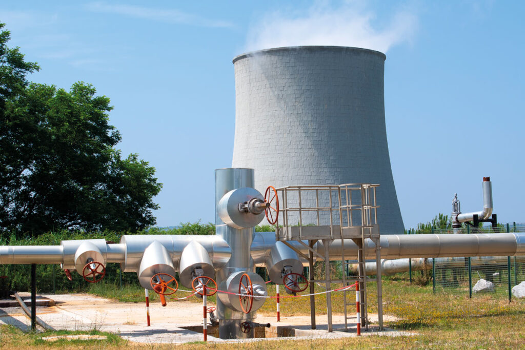 Im Hintergrund: Kühlturm eines Geothermiekraftwerks, Im Vordergrund: Ventilkonstruktion einer geothermischen Dampfleitung.