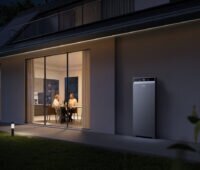 Blick von außen in ein hell erleuchetes Zimmer eines modernen Einfamilienhauses mit großer Fensterfront. Ein Mann und eine Frau sitzen an einem Tisch. Rechts neben dem Fenster steht auf der Terasse ein Speicher von Anker Solix.