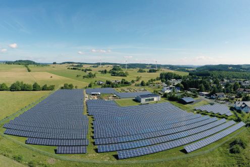 Blick auf einen Solarparkm im ländlichen Umfeld unter blauem Himmel. Im Hintergrund Windkraftanlagen.