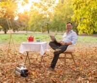 Mann an Tisch im Garten mit Laptop, roter Teekanne und mobilem Stromspeicher - Symbolbild für Axsol-Übernahme durch Loh.