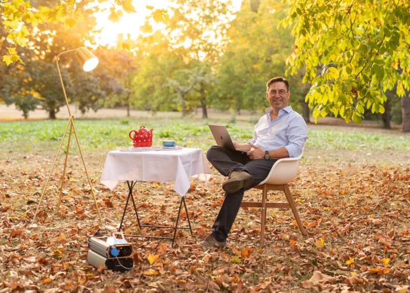 Mann an Tisch im Garten mit Laptop, roter Teekanne und mobilem Stromspeicher - Symbolbild für Axsol-Übernahme durch Loh.