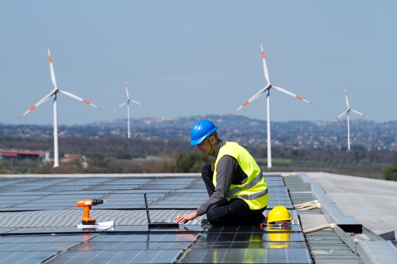Ein Handwerker montiert Solarmodule auf einem Flachdach. Im Hintergrund drehen sich SWindräder.