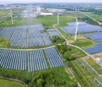 Im Vordergrund eine Photovoltaik-Freiflächenanlage mit Windkraftwerken, im Hintergrund ein Dorf.
