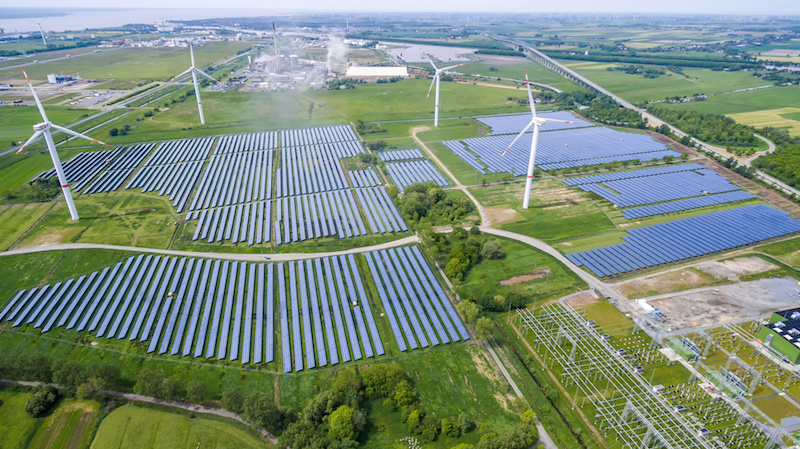 Im Vordergrund eine Photovoltaik-Freiflächenanlage mit Windkraftwerken, im Hintergrund ein Dorf.