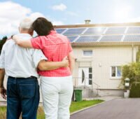Vorn ein älteres Paar, im Hintergrund ein Einfamilienhaus mit einer Photovoltaik-Anlage auf dem Dach