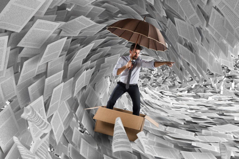 Ein Mann mit Krawatte und Regenschirm steht in einem Karton. Er befindet sich inmitten einer Papierflut. er navigiert durch diese Welle wie ein Wellensurfer.