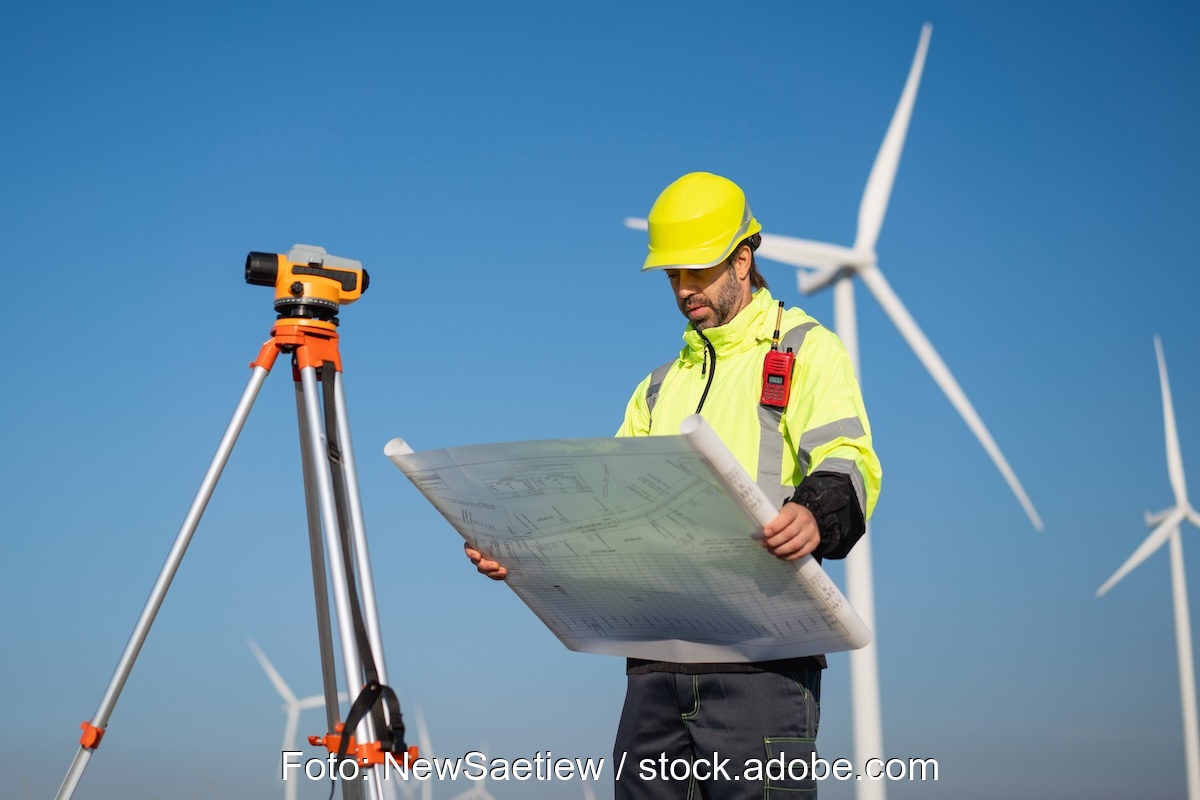 Das Foto zeigt einen Menschen in gelber Jacke mit gelber Kappe, der einen Plan in der Hand hält. Neben ihm ein Vermessungsgerät, im Hintergrund Windkraftanlagen (Beschleunigungsgebiete RED III)