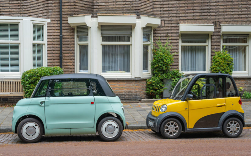 Ein türkisfarbenes und ein gelbes Kleinstfahrzeug parken hintereinander vor einem Backsteingebäude.