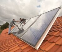 Im Vordergrund eine Solarthermie-Anlage auf einem Dach. Sie wird von zwei Handwerkern installiert.