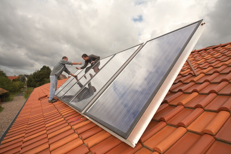 Im Vordergrund eine Solarthermie-Anlage auf einem Dach. Sie wird von zwei Handwerkern installiert.