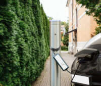 Ladesäule für ein Elektroauto neben einem Busch, im Hintergrund ein Haus. Angeschlossen ist ein Fahrzeug.