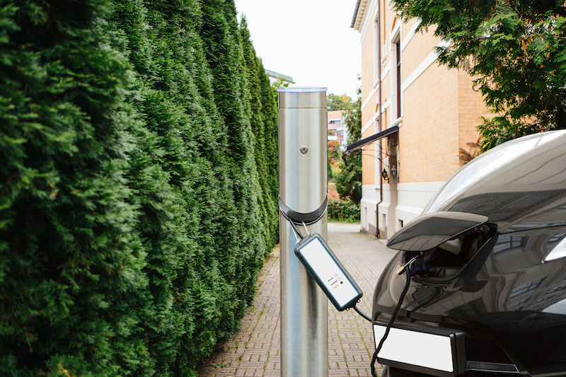 Ladesäule für ein Elektroauto neben einem Busch, im Hintergrund ein Haus. Angeschlossen ist ein Fahrzeug.