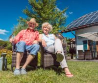 Ein älteres Paar sitzt in seinem Garten, im Hintergrund ein Haus mit einer kleinen PV-Anlage auf dem Dach.