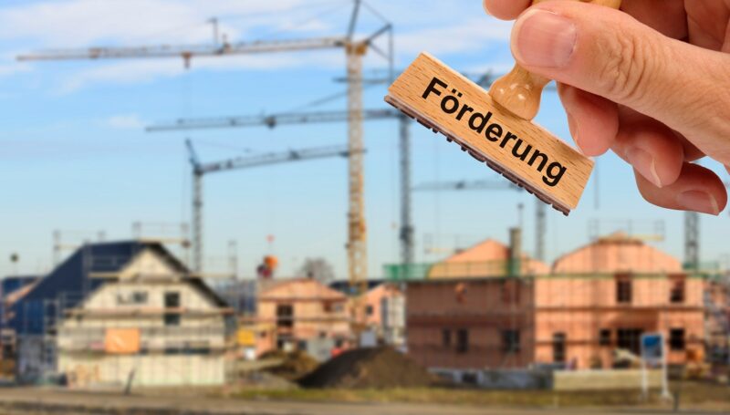 Häuser im Bau und Hand mit Stempel Förderung, die progres.nrw-Förderprogramme für Elektromobilität und Klimaschutztechnik werden fortgesetzt.