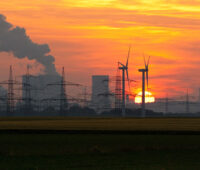 Vor einem orangen Abendhimmel mit untergehender Sonne sind die Kühltürme eines Kohlekraftwerks zu sehen, davor Strommasten und Windkraftanlagen.