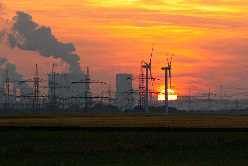 Vor einem orangen Abendhimmel mit untergehender Sonne sind die Kühltürme eines Kohlekraftwerks zu sehen, davor Strommasten und Windkraftanlagen.