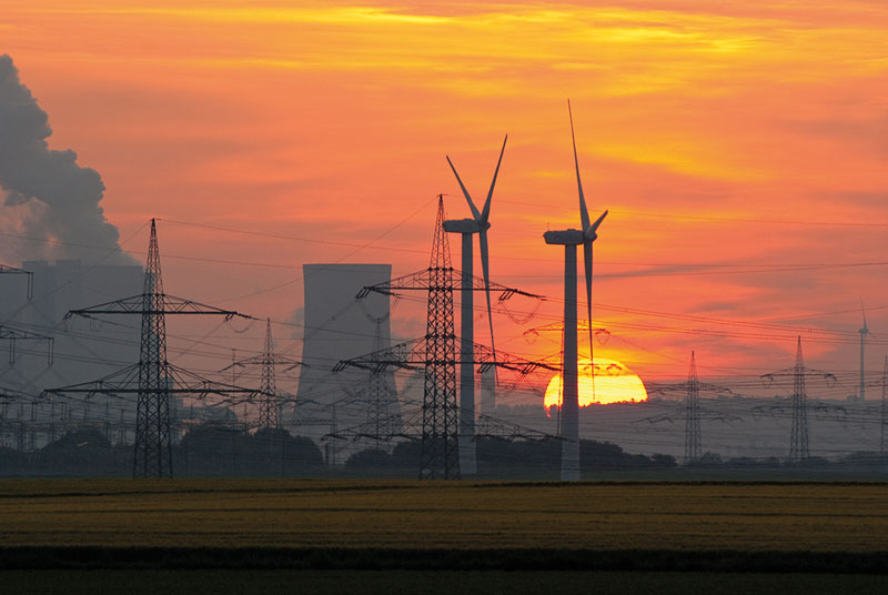 Energiewende. Windkraftanlagen vor Kohlekraftwerk und Strommasten. Ganz im Hintergrund die untergehende Sonne.