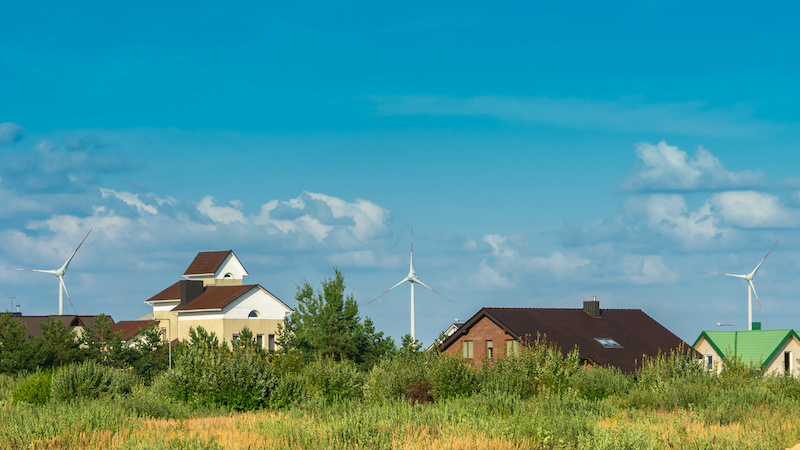 Im Vordergrund eine Wiese und Gebäude, im Hintergrund Windkraftanlagen