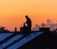 Schornsteinfeger im Abendrot auf einem Dach beim Reinigen des Schornsteins, auf Dach auch eine Solaranlage, Ziel ist ein 65-Prozent-Anteil erneuerbarer Energie