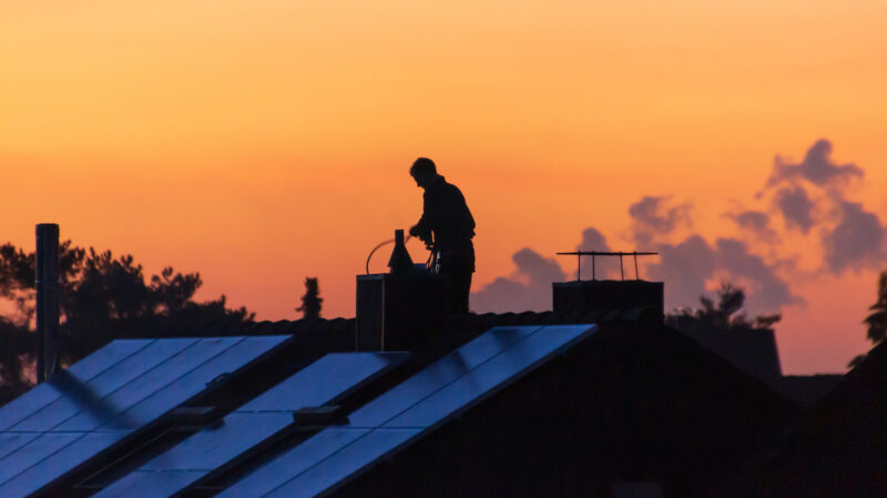 Schornsteinfeger im Abendrot auf einem Dach beim Reinigen des Schornsteins, auf Dach auch eine Solaranlage, Ziel ist ein 65-Prozent-Anteil erneuerbarer Energie