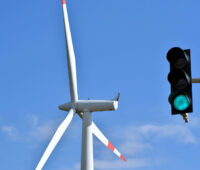 Im Vordergrund vor blauem Himmel eine grün zeigende Ampel, im Hintergrund eine Windkraftanlage