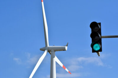 Im Vordergrund vor blauem Himmel eine grün zeigende Ampel, im Hintergrund eine Windkraftanlage