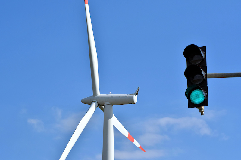 Im Vordergrund vor blauem Himmel eine grün zeigende Ampel, im Hintergrund eine Windkraftanlage