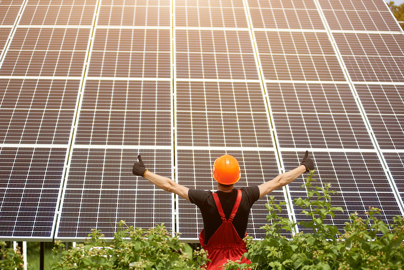 Ein Arbeiter mit orangem Helm und roter Latzhose steht vor Photovoltaik-Anlage, streckt die Arme auseinander und zeit mit en Daumen nach oben.