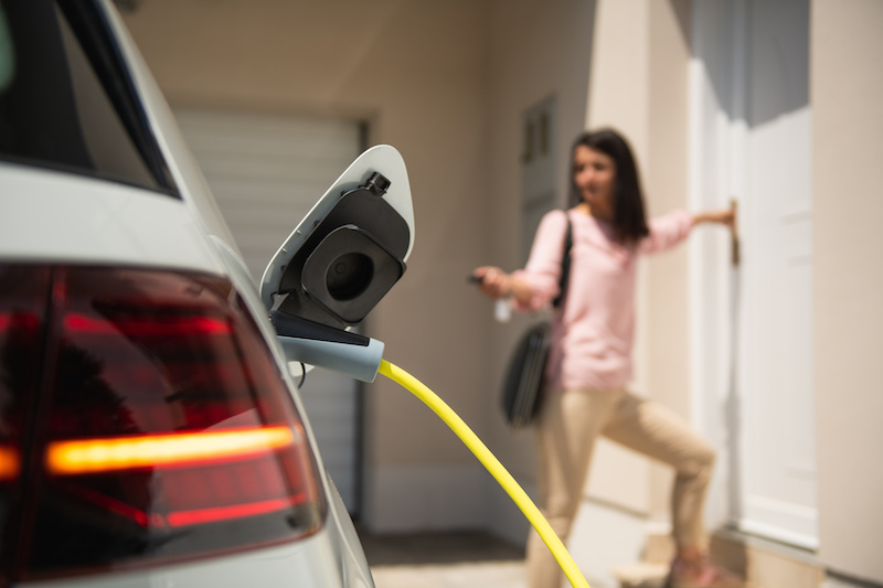 im Vordergrund ein Elektrofahrzeug, an das ein Ladekabel angeschlossen ist. Im Hintergrund eine Frau, die eine Haustür öffnet.