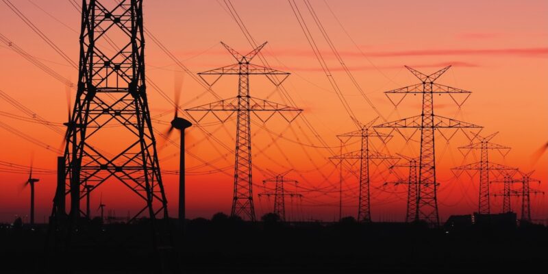 Strommasten von Hochspannungsleitungen bei Sonnenuntergang mit Windrädern.