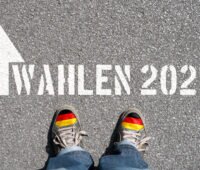 Straßenbelag, auf den "Wahlen 2021" geschrieben wurde. Davor unten im Bild ein paar Schuhe. Bald ist Wahlkampf.