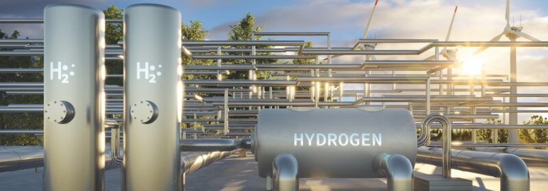 Im Vordergrund Speichertanks für Hydrogen (Wasserstoff), im Hintergrund Röhren sowie dahinter Windkraftanlagen für die Produktion von grünem Wasserstoff.